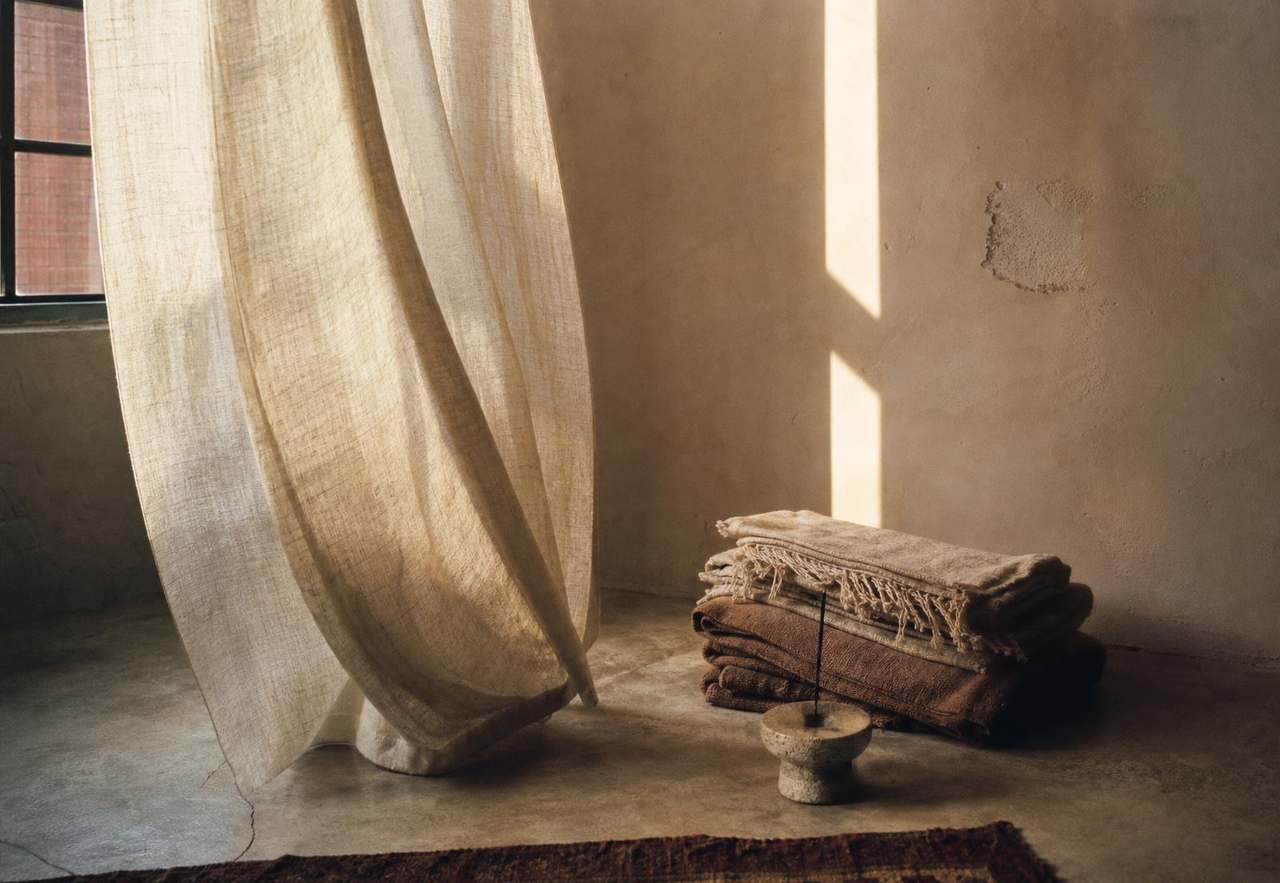 Afternoon light on a plaster wall beside folded blankets, linen curtain and a stone incense holder at the SACRAHAUS New York space